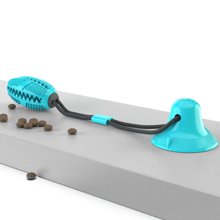 A turquoise and black suction cup dog toy with an oval-shaped spiked ball is attached to a flat surface. Small pieces of dog kibble are scattered around the toy.