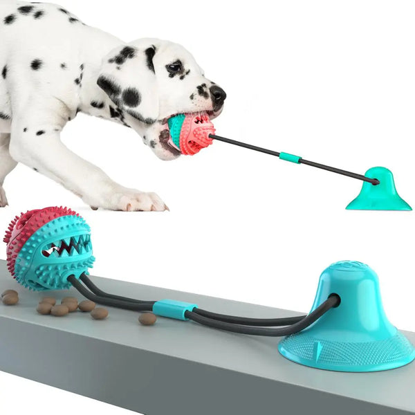 A Dalmatian puppy is enthusiastically tugging on a suction cup dog toy with a spiked, multicolored ball. The toy is securely attached to a flat surface, and a section of dog kibble is scattered near the toy.