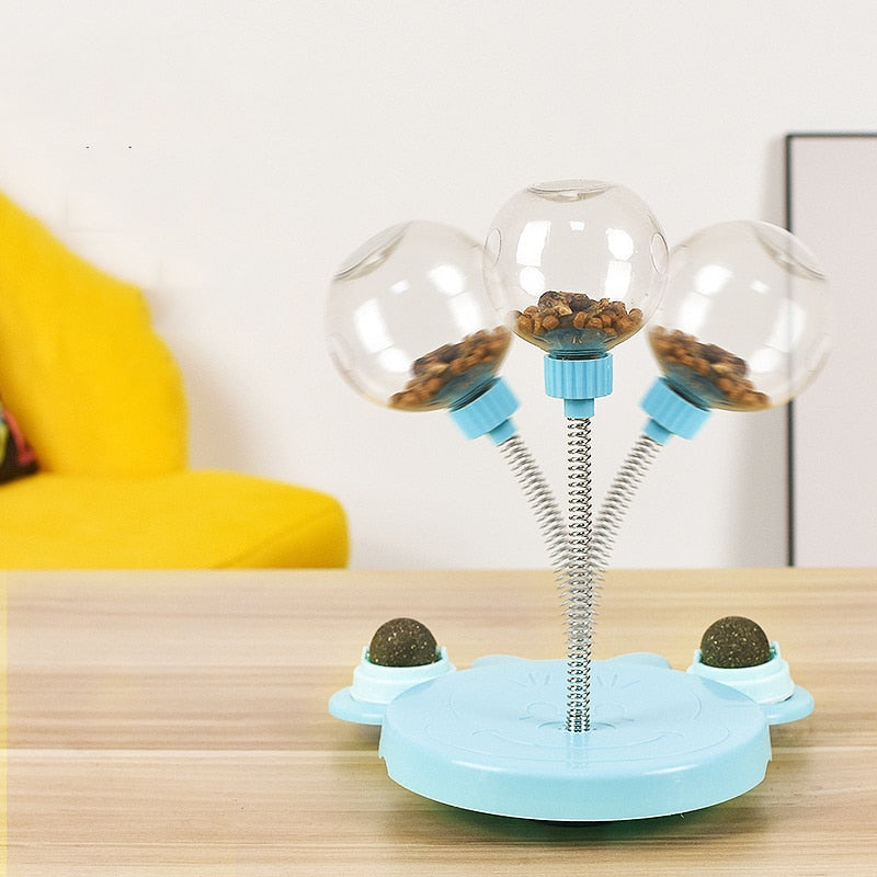 A blue interactive cat toy placed on a wooden table, featuring three spring-mounted clear balls filled with treats. The toy also includes two side compartments with treat balls, designed to keep cats entertained and engaged.