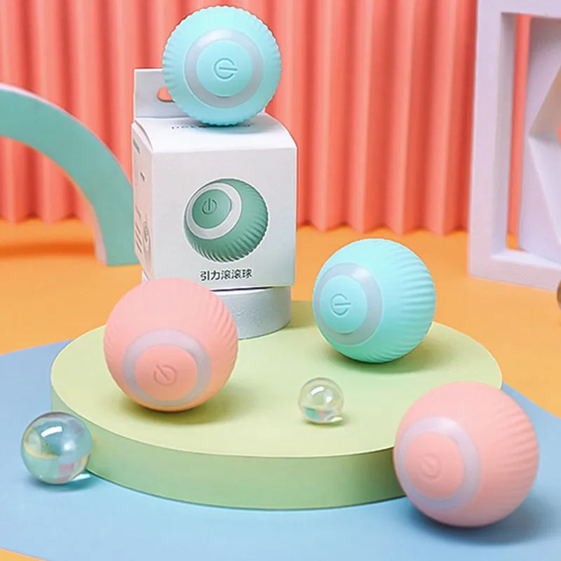 A display of multiple pastel-colored automatic cat teaser balls, including teal and coral variants. The balls are arranged around a packaging box, emphasizing their appealing design and playful functionality.