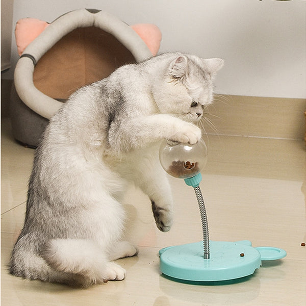 A gray and white cat interacts with a blue interactive treat-dispensing toy. The toy has a spring-mounted clear ball filled with treats and a sturdy base, engaging the cat in playful activity.